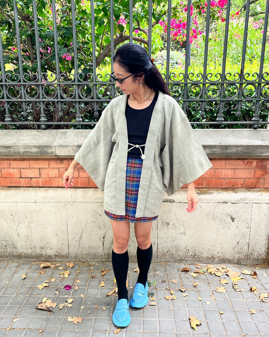 A woman wearing a gray haori jacket over a black top, with a multicolored skirt and blue shoes, standing by a gate with green plants in the background.