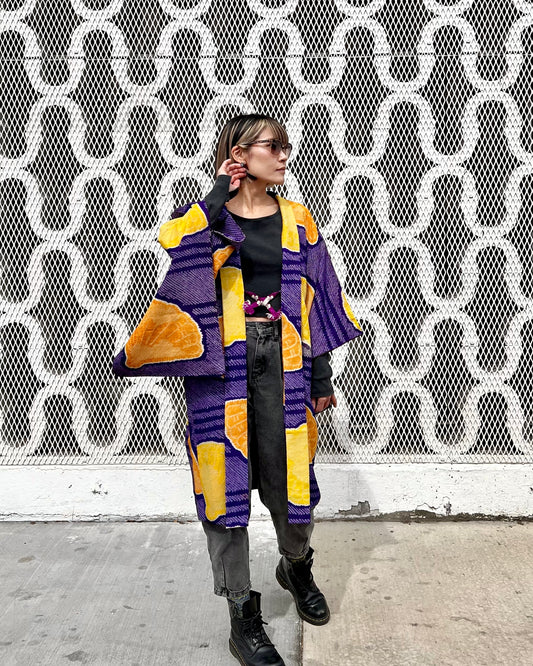 A person wearing a vibrant purple and yellow shibori-dyed haori with a floral and crane printed lining, standing in front of a wall with a geometric pattern.