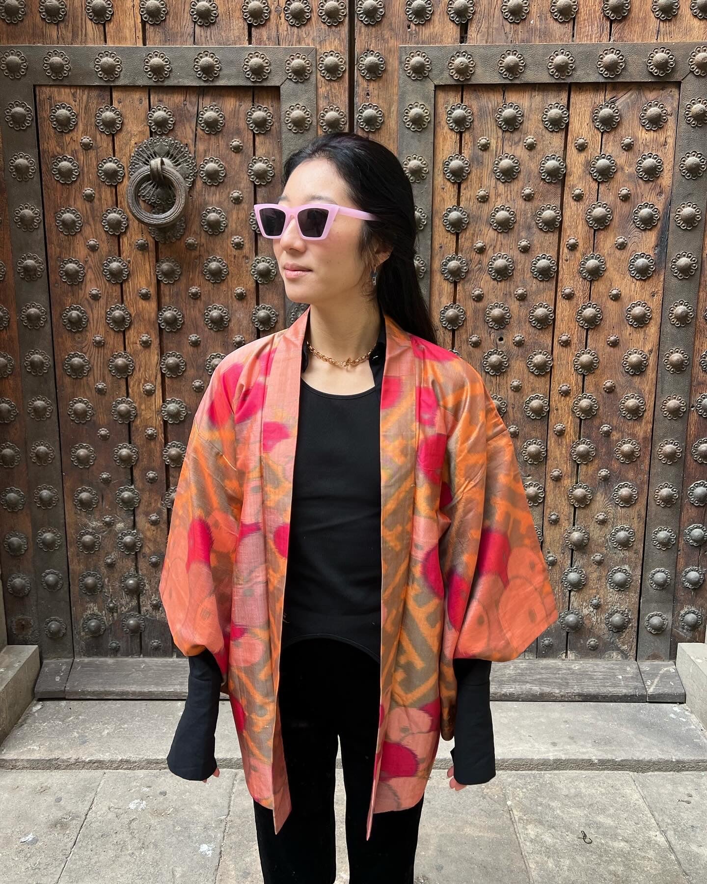 A woman standing in front of a traditional wooden door wearing a multicolored antique haori jacket with a geometric pattern.